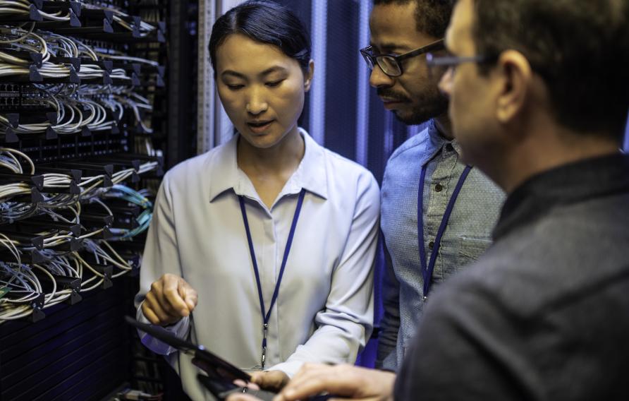 Three IT technician looking at a digital tablet and talking while standing next to a server in a data center.